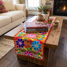 Load image into Gallery viewer, A colorful embroidered table runner with red pom-pom trim draped over a wooden coffee table in a living room setting. The runner features vibrant floral and geometric patterns in pink, blue, yellow, green, and orange.
